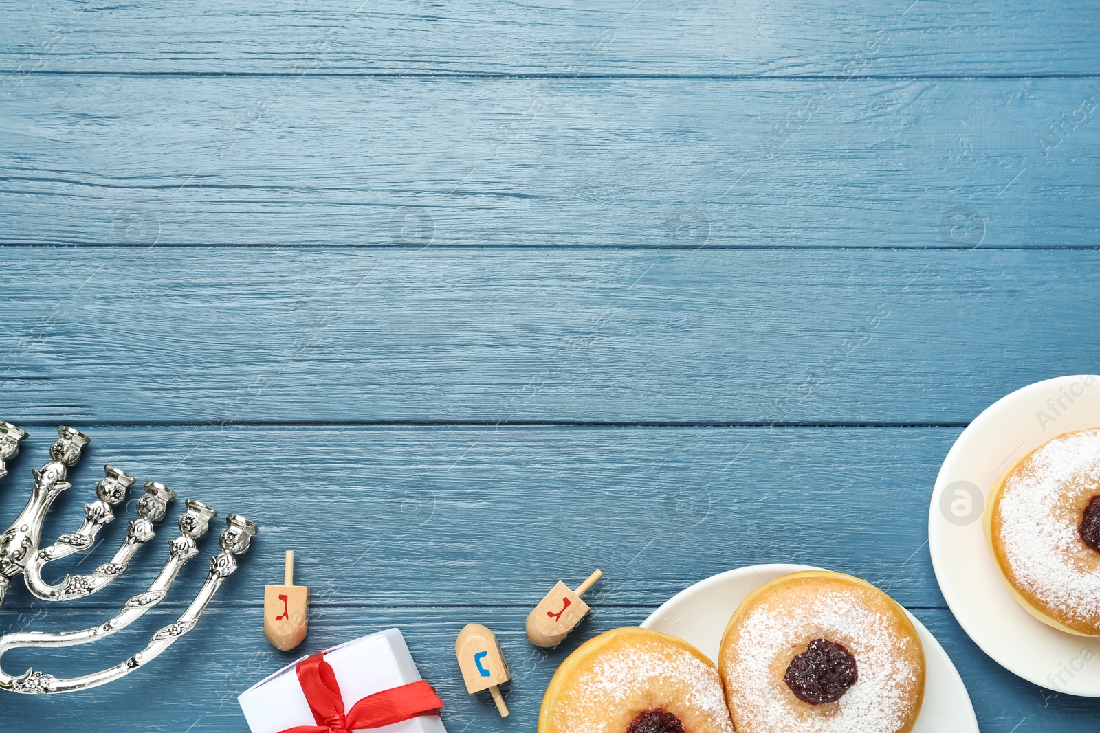 Photo of Flat lay composition with menorah, gift box, dreidels icluding He, Pe, Nun, Gimel letters and sufganiyot on blue wooden table, space for text. Hanukkah symbols