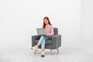 Happy young woman with laptop sitting in armchair near white wall indoors