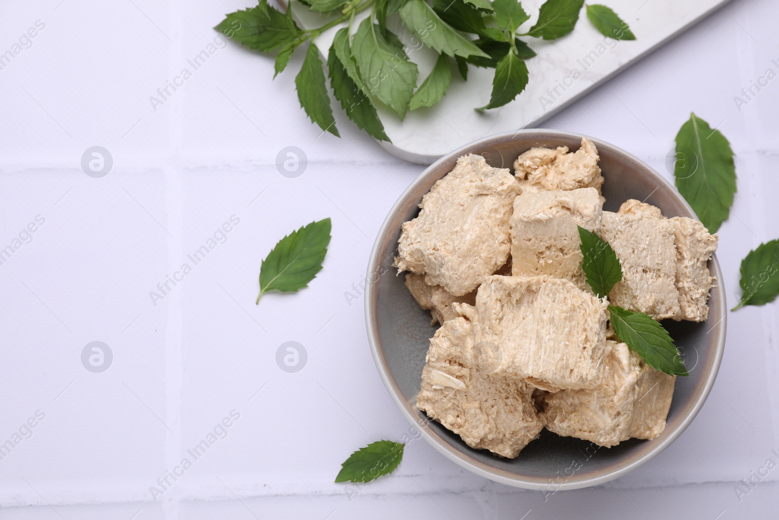 Photo of Bowl with pieces of tasty halva and mint leaves on white tiled table, flat lay. Space for text