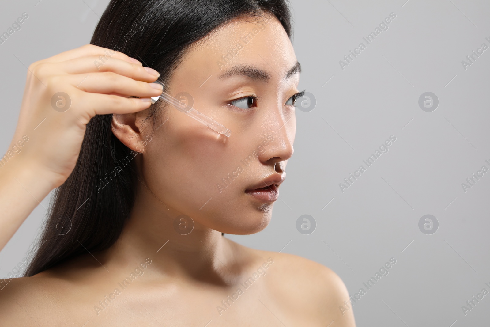 Photo of Beautiful young woman applying cosmetic serum onto her face on grey background, space for text