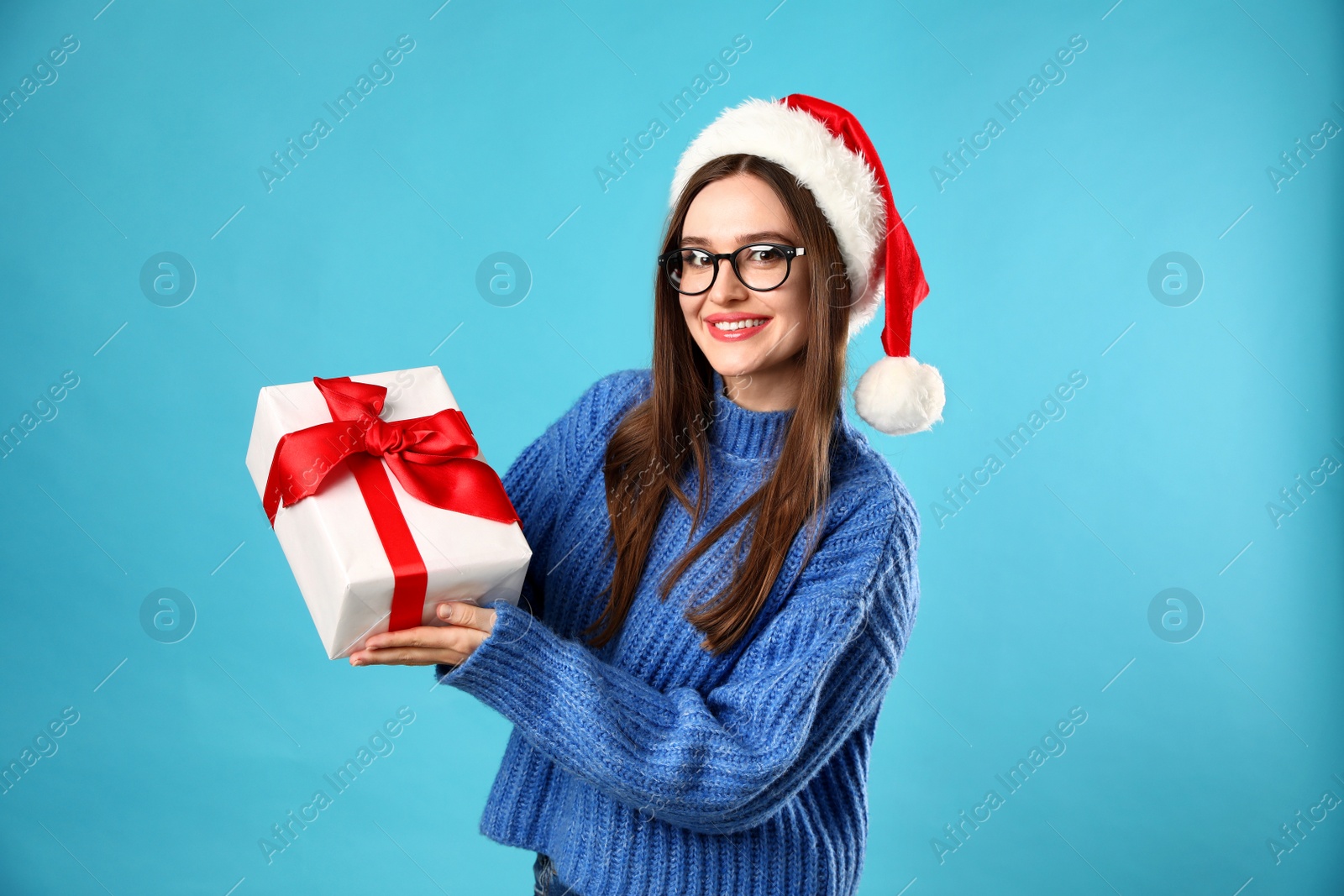 Photo of Young woman in Christmas sweater with Santa hat and gift box on blue background