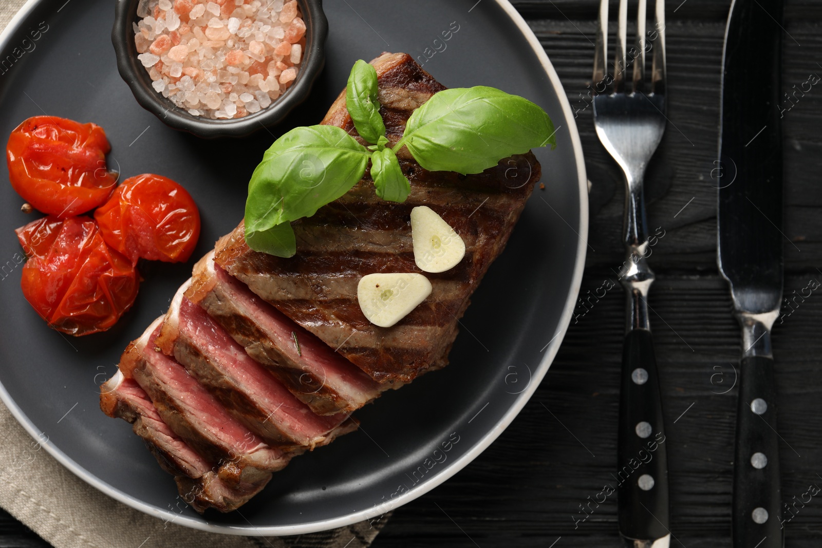 Photo of Delicious grilled beef steak served with spices and tomatoes on black wooden table, flat lay