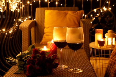 Photo of Bouquet of roses, glasses with wine and burning candles on rattan table at balcony in night