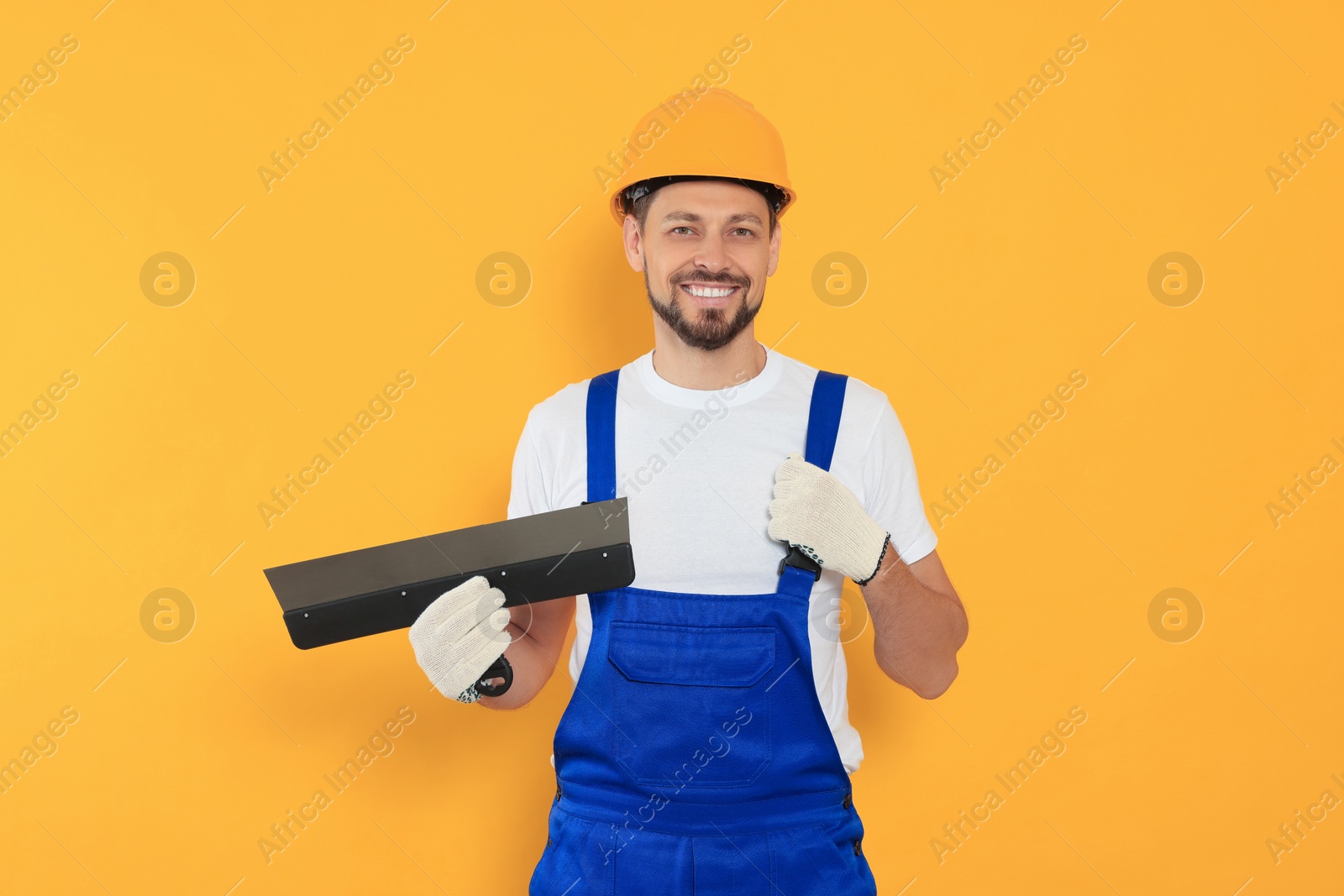 Photo of Professional worker with putty knife in hard hat on orange background