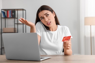 Photo of Confused woman with credit card talking on phone in front of laptop at table indoors. Be careful - fraud