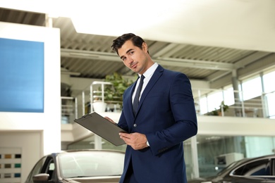 Young salesman with clipboard near car in modern dealership