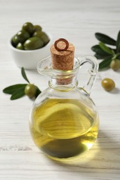 Photo of Glass jug of oil, ripe olives and green leaves on white wooden table