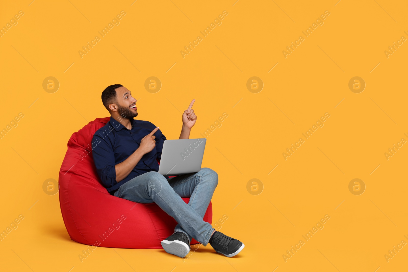 Photo of Smiling young man with laptop sitting on beanbag chair against yellow background, space for text
