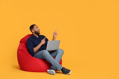 Smiling young man with laptop sitting on beanbag chair against yellow background, space for text