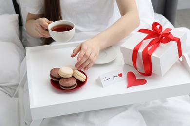 Photo of Tasty breakfast served in bed. Woman with tea, macarons, gift box and I Love You card at home, closeup