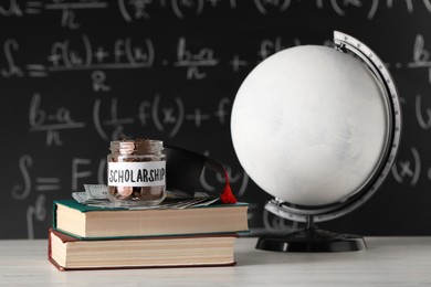 Photo of Scholarship concept. Globe, glass jar with coins, graduation cap, dollar banknotes and books on white wooden table
