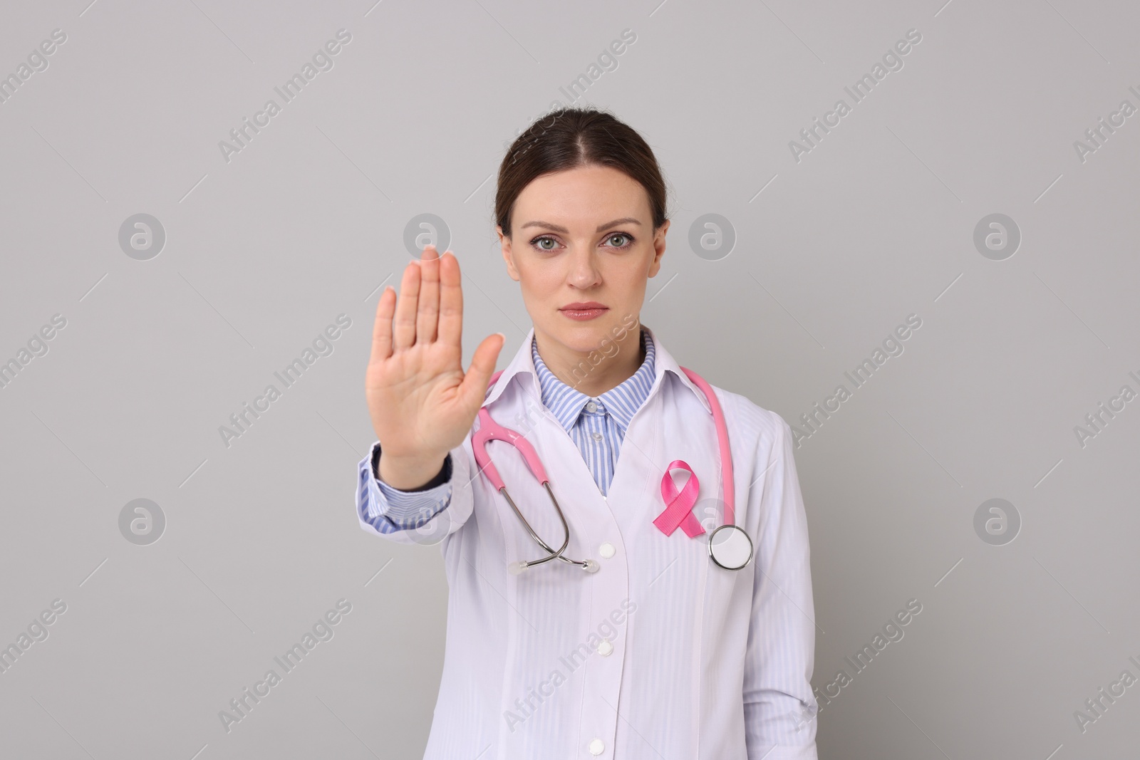 Photo of Mammologist with pink ribbon showing stop gesture against grey background. Breast cancer awareness