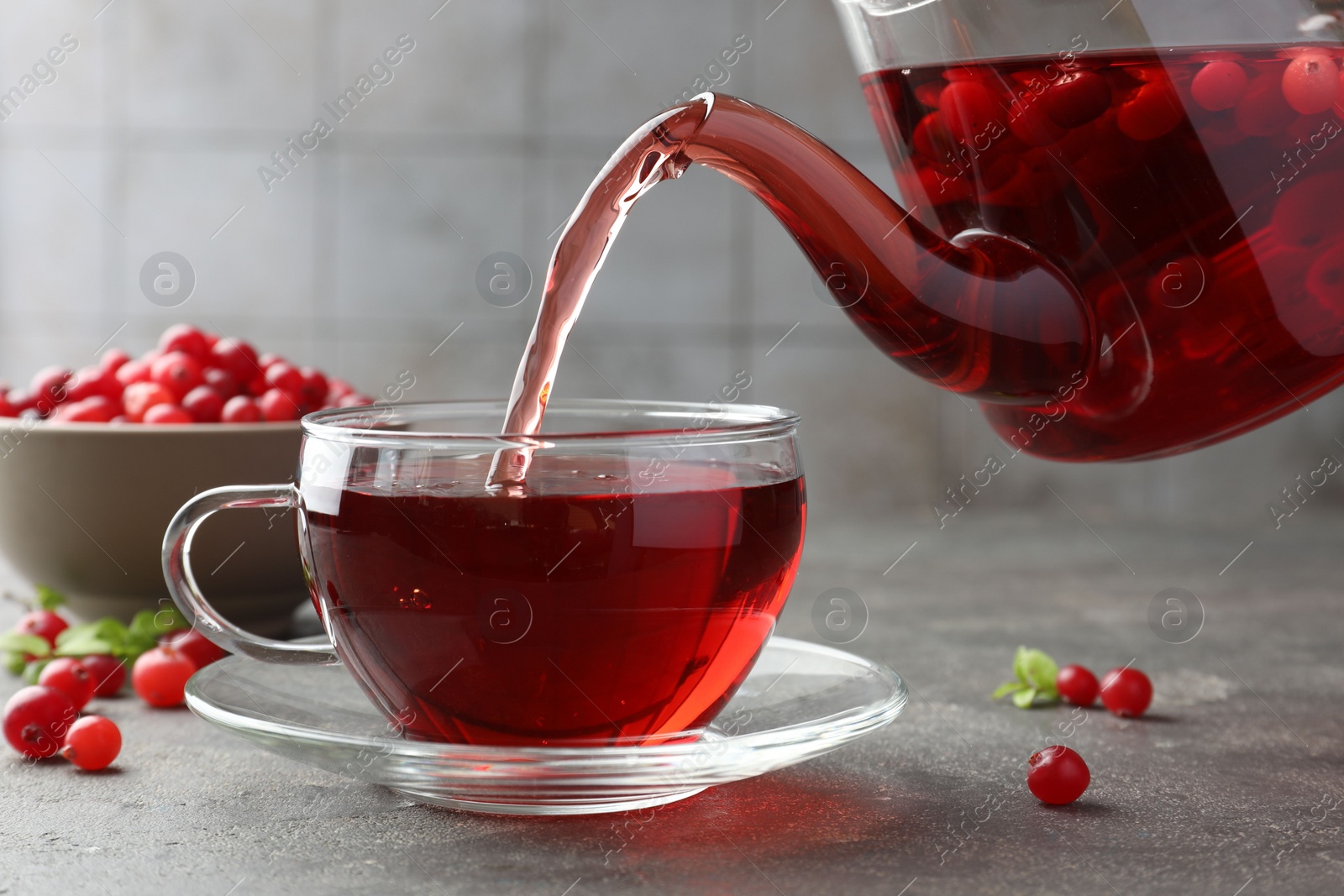 Photo of Pouring hot cranberry tea into glass cup and fresh berries on light grey textured table, closeup