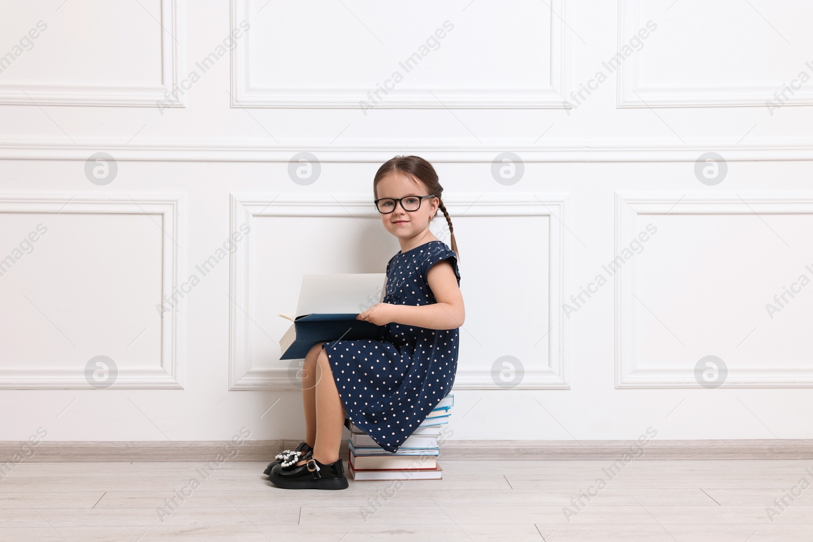 Photo of Cute little girl in glasses sitting on stack of books near white wall