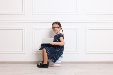 Cute little girl in glasses sitting on stack of books near white wall