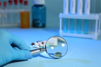 Scientist with magnifying glass examining pills at light blue table, closeup