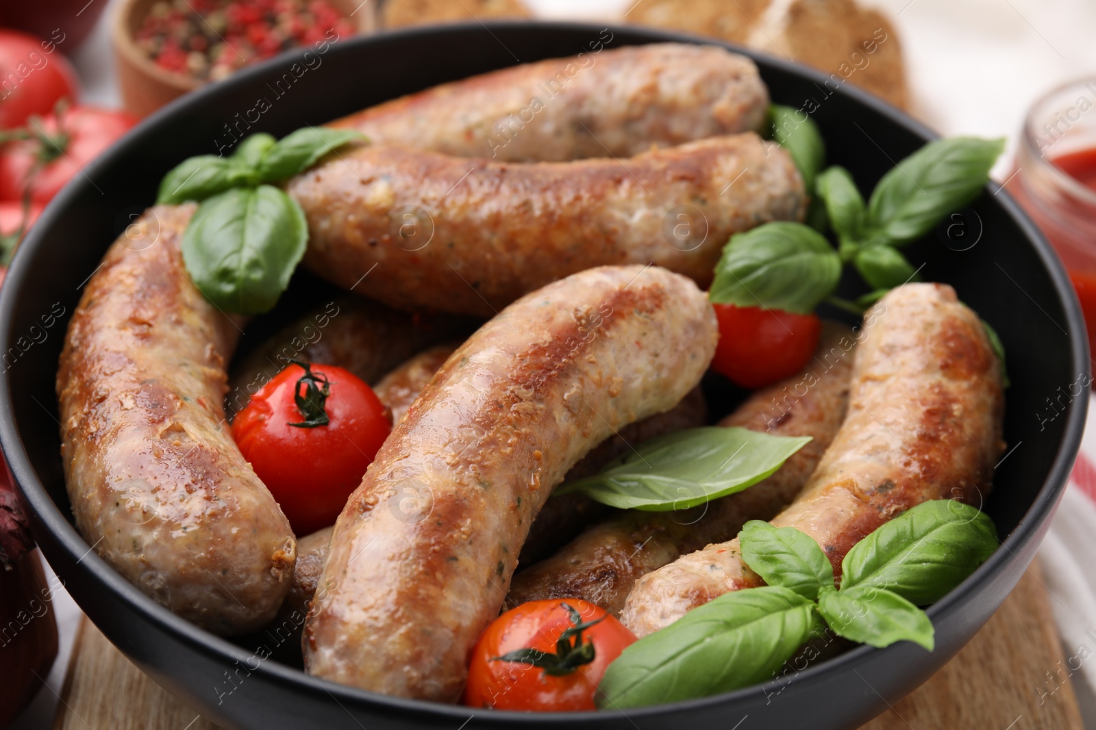 Photo of Bowl with tasty homemade sausages, basil leaves and tomatoes on table, closeup