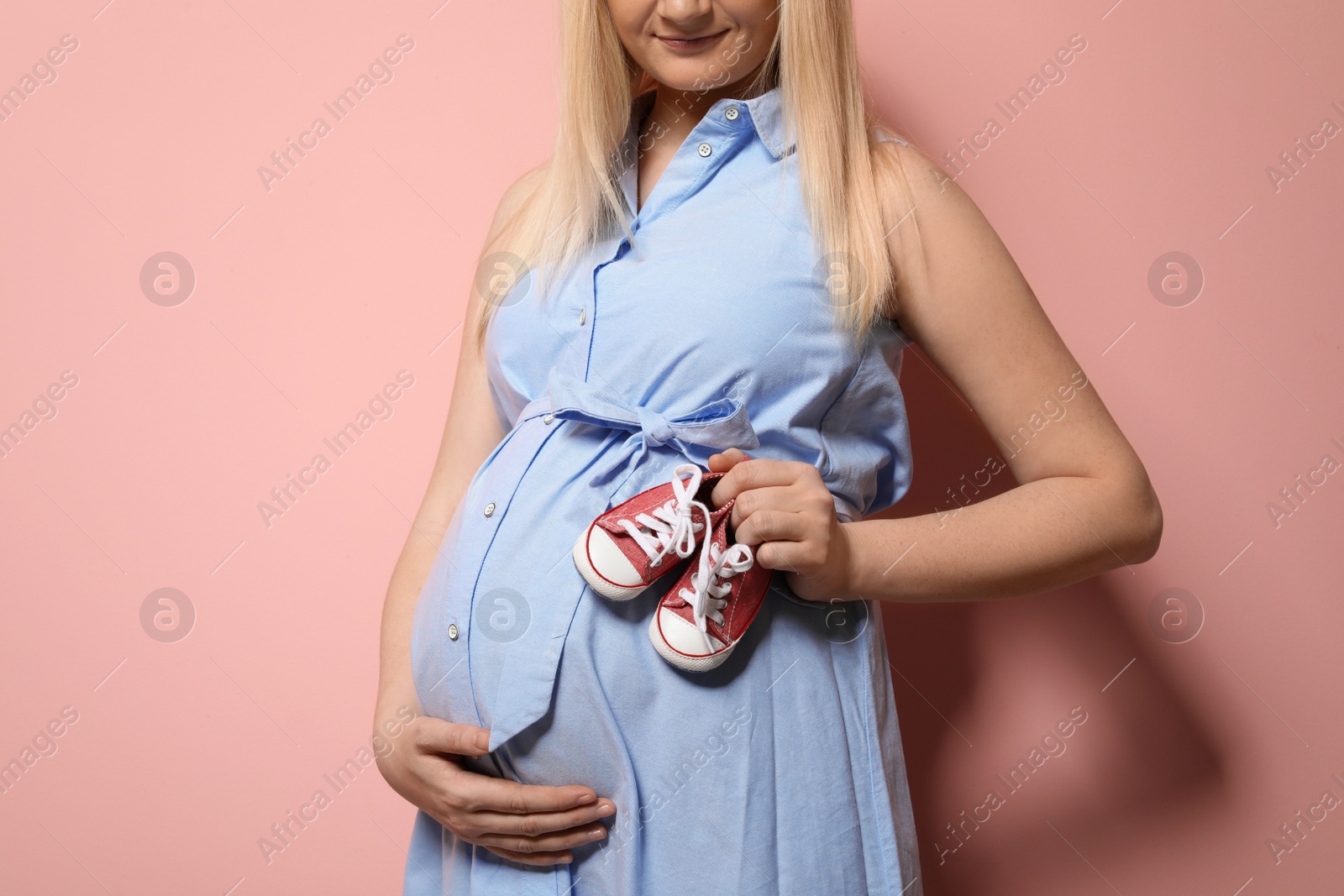 Photo of Pregnant woman in dress holding baby booties near tummy on color background