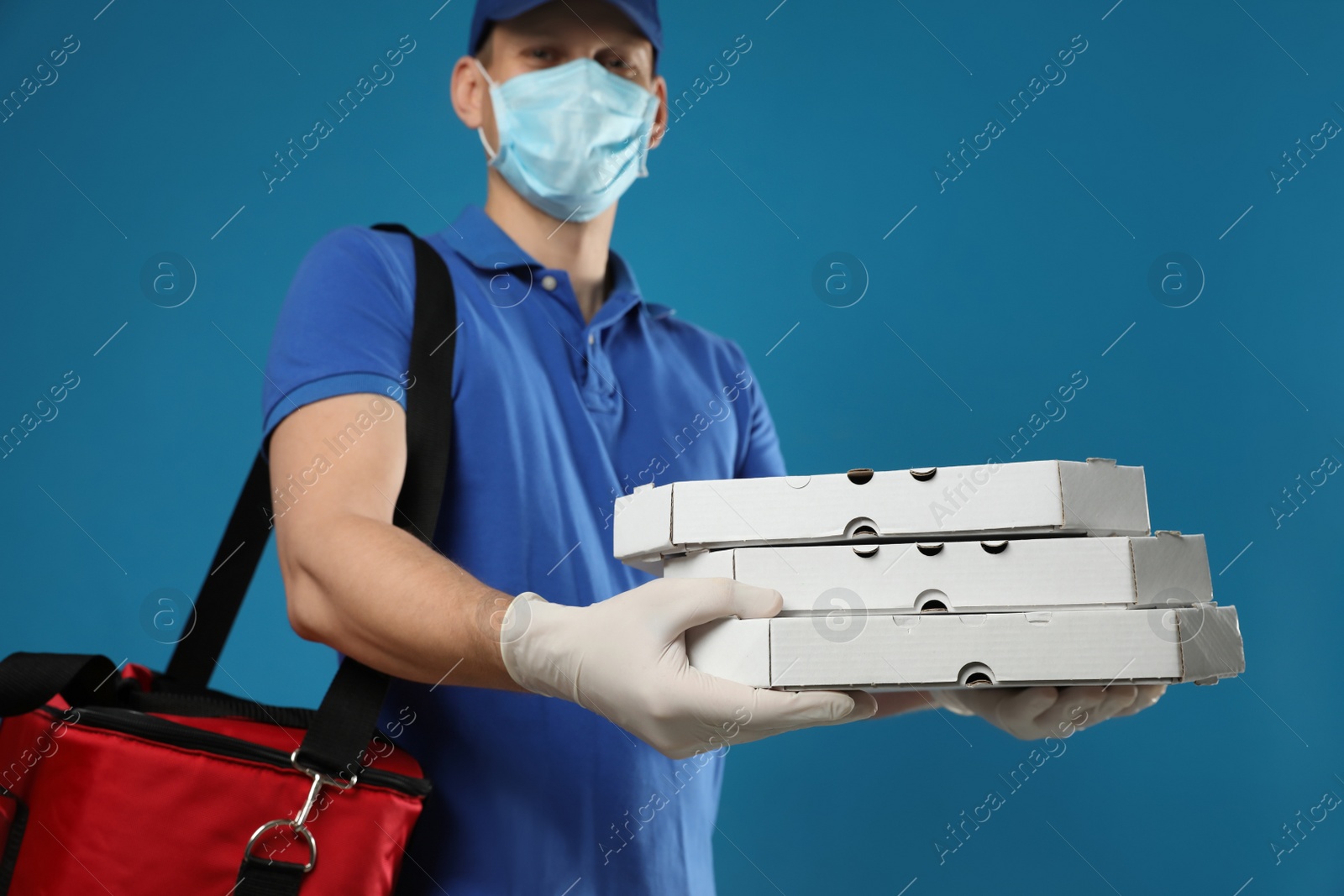 Photo of Courier in protective mask and gloves holding pizza boxes on blue background. Food delivery service during coronavirus quarantine