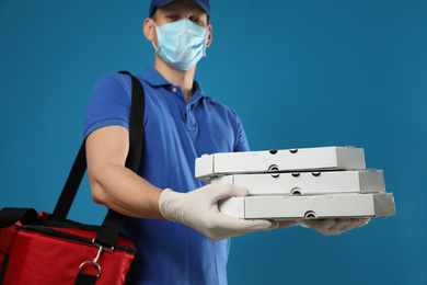 Courier in protective mask and gloves holding pizza boxes on blue background. Food delivery service during coronavirus quarantine