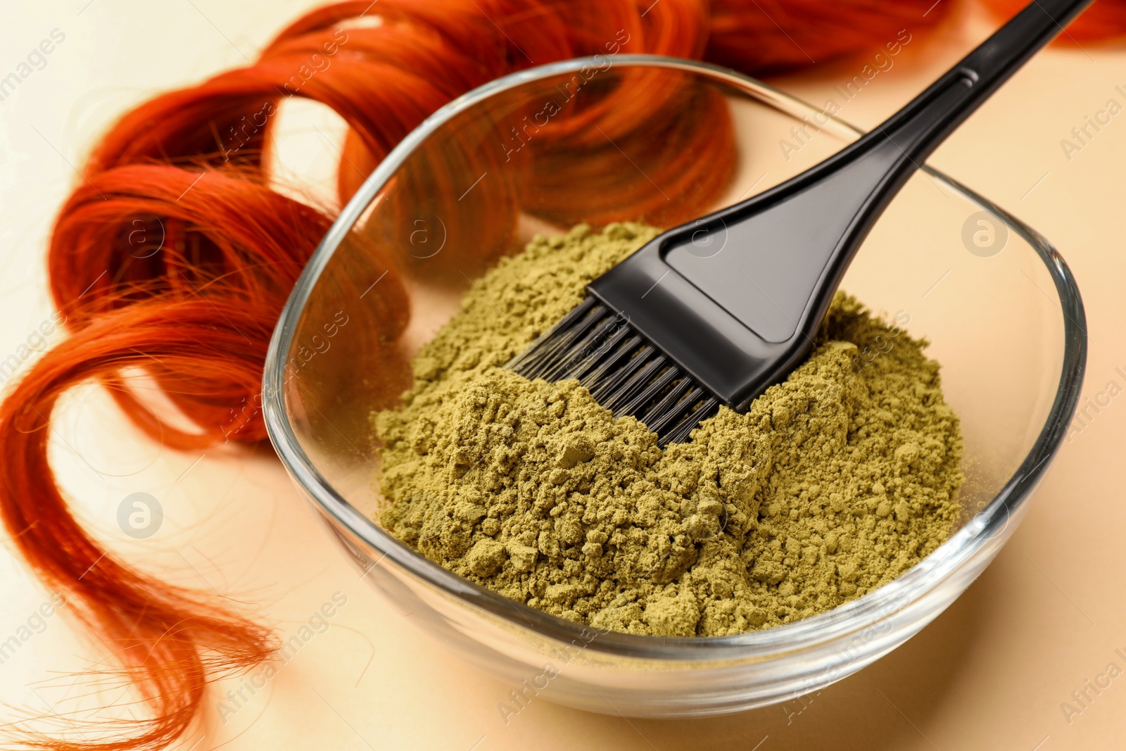 Photo of Bowl of henna powder, brush and red strand on beige background, closeup. Natural hair coloring