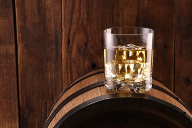 Whiskey with ice cubes in glass on barrel against wooden background, space for text