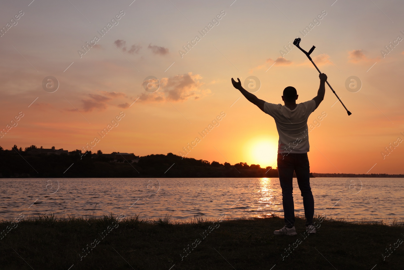 Photo of Man raising elbow crutch up to sky near river at sunset, back view. Healing miracle