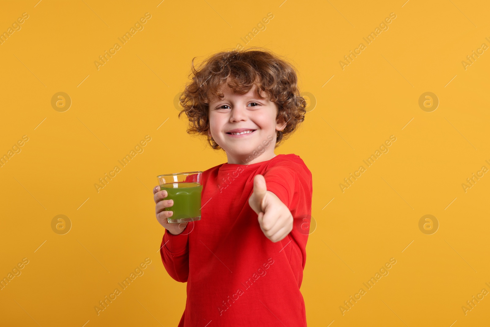 Photo of Cute little boy with glass of fresh juice showing thumb up on orange background