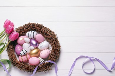 Photo of Many painted Easter eggs, tulip flowers and ribbon on white wooden table, flat lay. Space for text