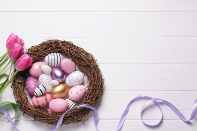 Many painted Easter eggs, tulip flowers and ribbon on white wooden table, flat lay. Space for text