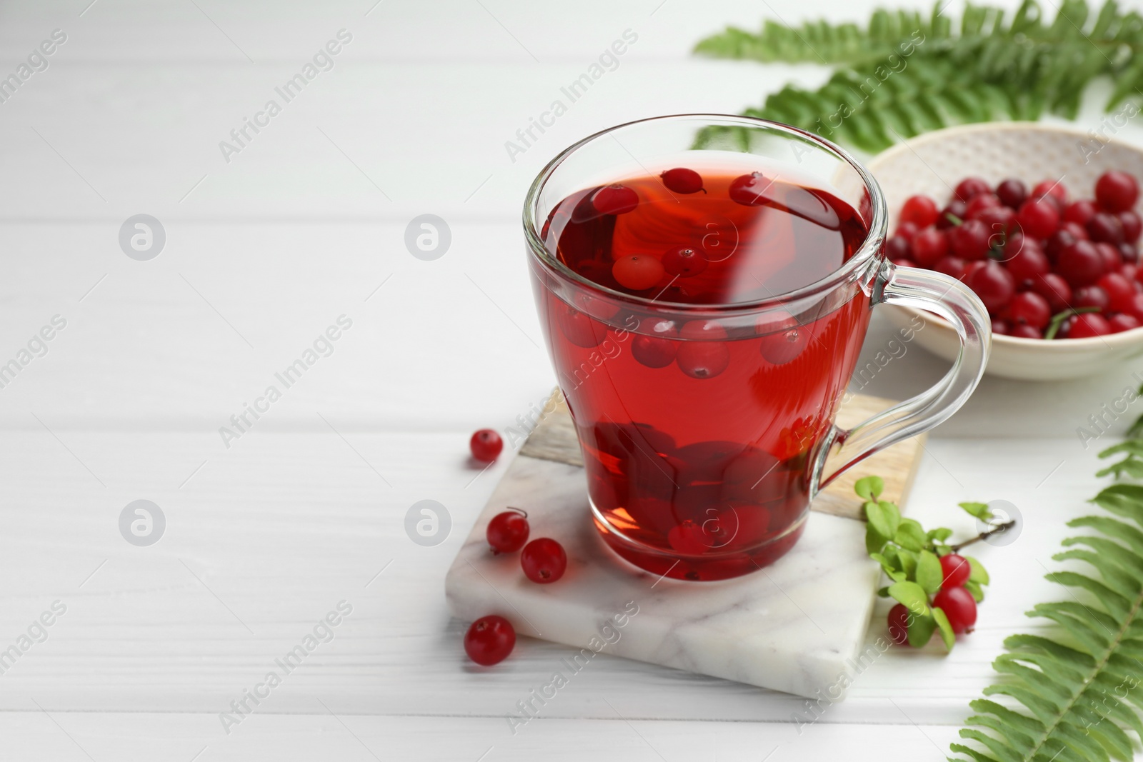 Photo of Tasty hot cranberry tea in glass cup, fresh berries and leaves on white wooden table