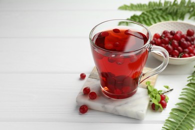 Tasty hot cranberry tea in glass cup, fresh berries and leaves on white wooden table