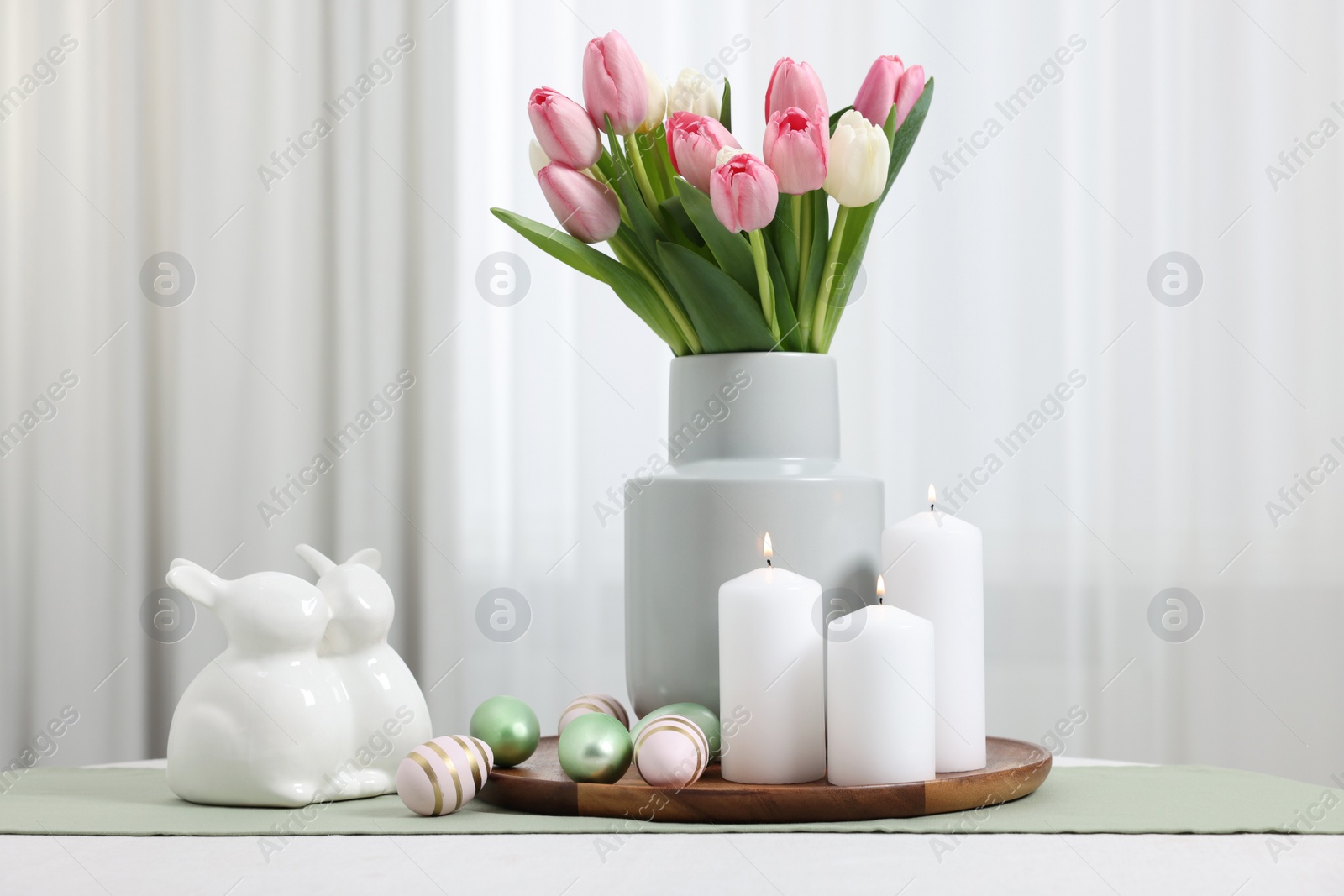 Photo of Easter decorations. Bouquet of tulips, painted eggs, burning candles and bunny figures on white table indoors