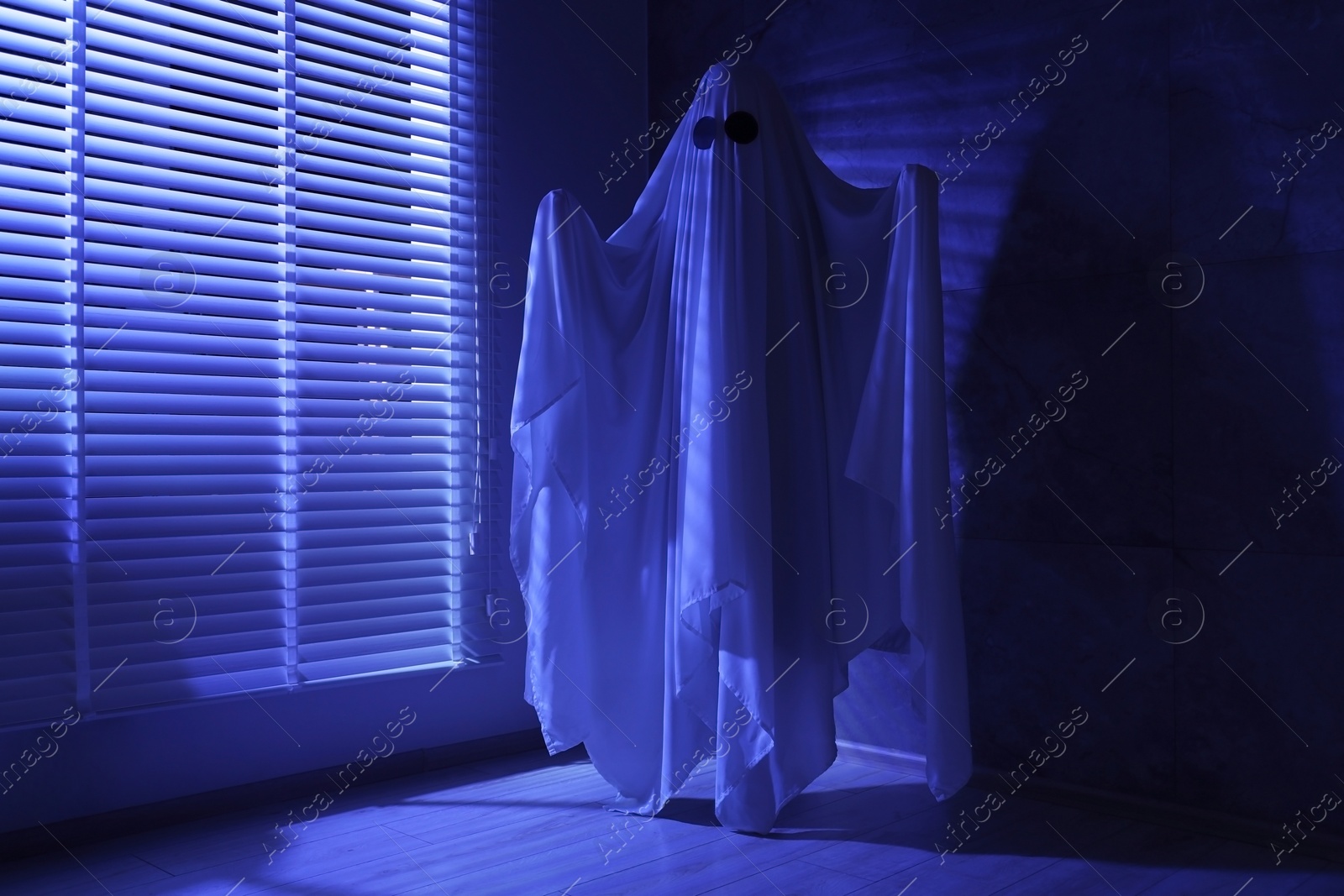 Photo of Creepy ghost. Woman covered with sheet near window in blue light