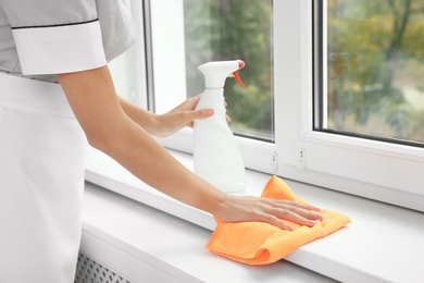Photo of Young chambermaid cleaning window sill with rag indoors, closeup