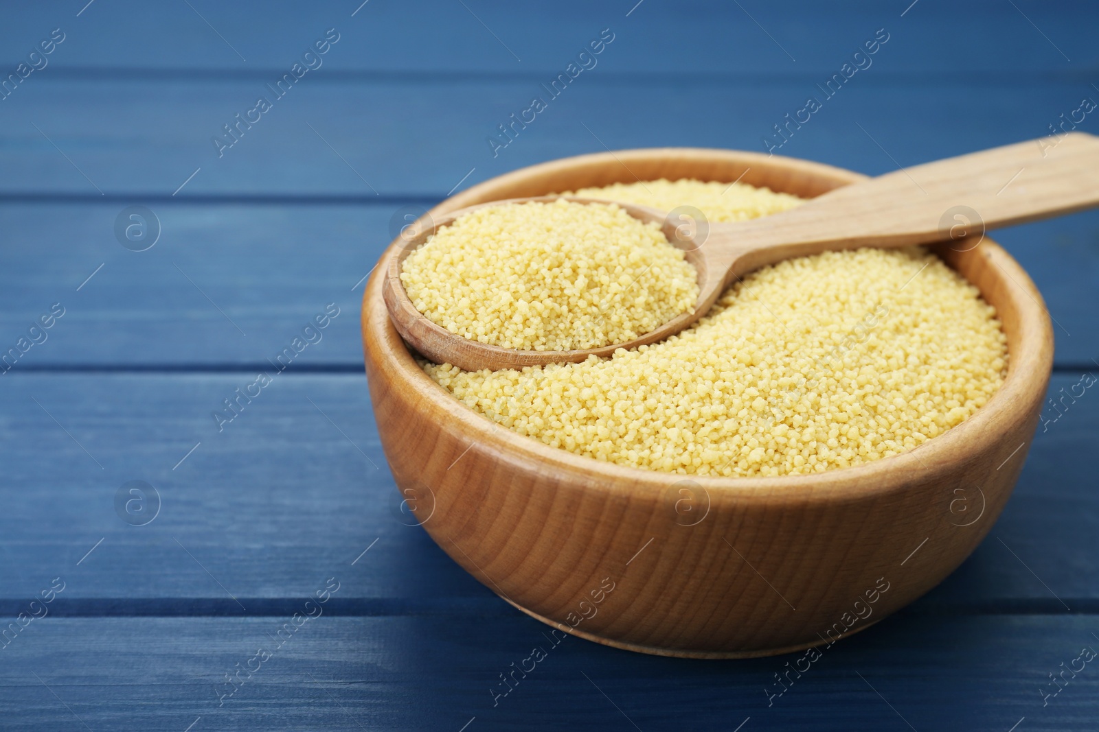 Photo of Bowl and spoon with raw couscous on blue wooden table, closeup. Space for text