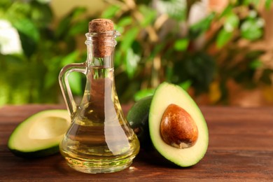 Photo of Glass jug of cooking oil and fresh avocados on wooden table against blurred green background