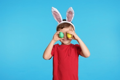 Little boy in bunny ears headband holding Easter eggs near eyes on color background