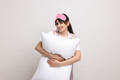 Happy woman in pyjama and sleep mask holding pillow on light grey background