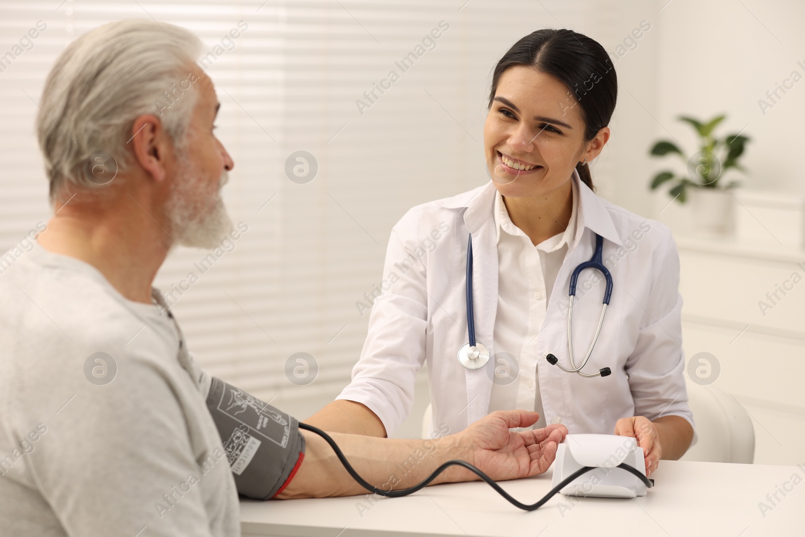 Photo of Smiling nurse measuring elderly patient's blood pressure at white table in hospital