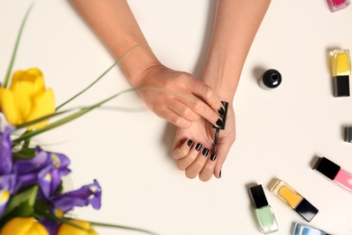 Photo of Woman applying nail polish near bottles on white table with flowers, top view