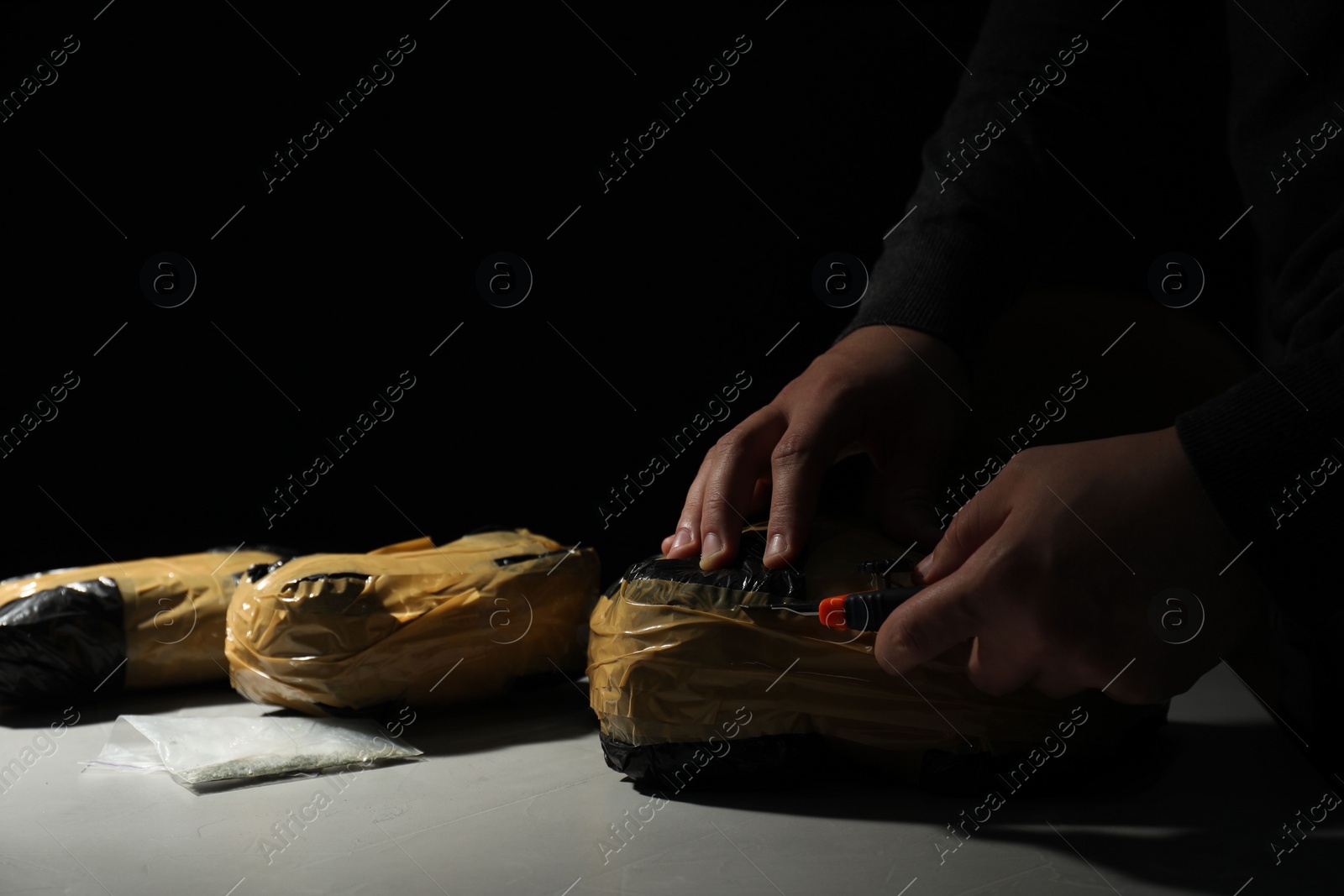 Photo of Smuggling and drug trafficking. Man opening package of narcotics with box cutter at light table against black background, closeup. Space for text