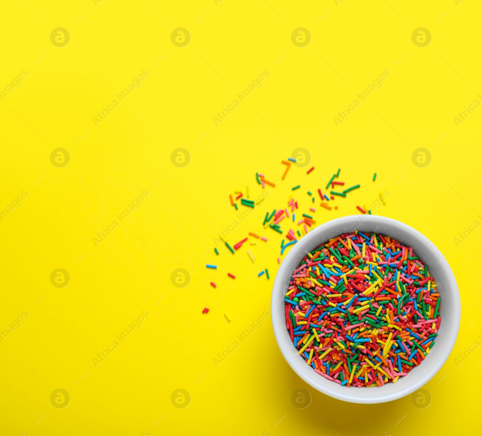 Photo of Colorful sprinkles in bowl on yellow background, top view with space for text. Confectionery decor