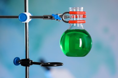 Photo of Flask with green liquid on retort stand against blurred background, closeup