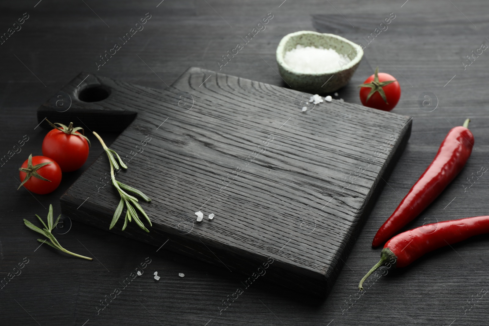 Photo of Cutting board, rosemary, salt, chili peppers and tomatoes on black wooden table. Space for text