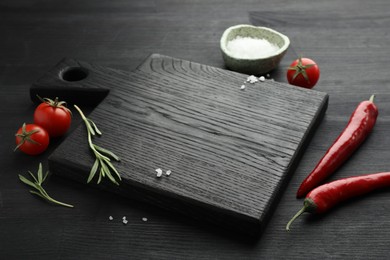 Photo of Cutting board, rosemary, salt, chili peppers and tomatoes on black wooden table. Space for text