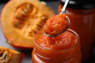 Photo of Taking delicious pumpkin jam from jar with spoon near fresh pumpkin on grey table, closeup