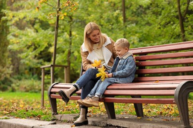 Happy mother and her son spending time together with dry leaves on wooden bench in autumn park