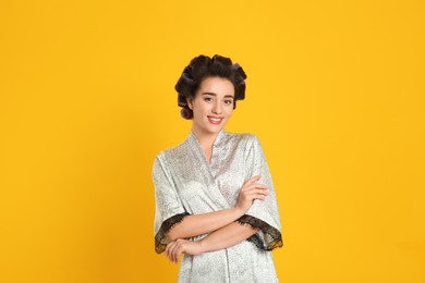 Photo of Happy young woman in silk bathrobe with hair curlers on orange background