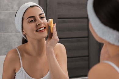 Photo of Young woman with headband washing her face using sponge near mirror in bathroom